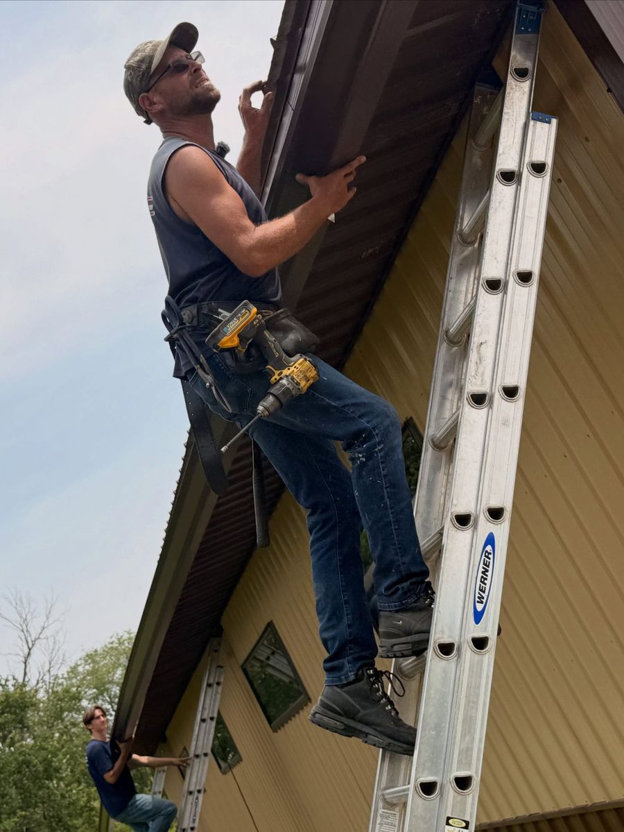 Installer on a ladder fitting a dark brown gutter under the eave of a tan steel-sided building