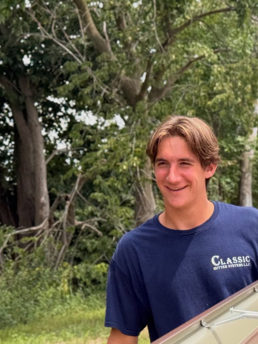 A smiling young crew member carrying a length of gutter under the trees