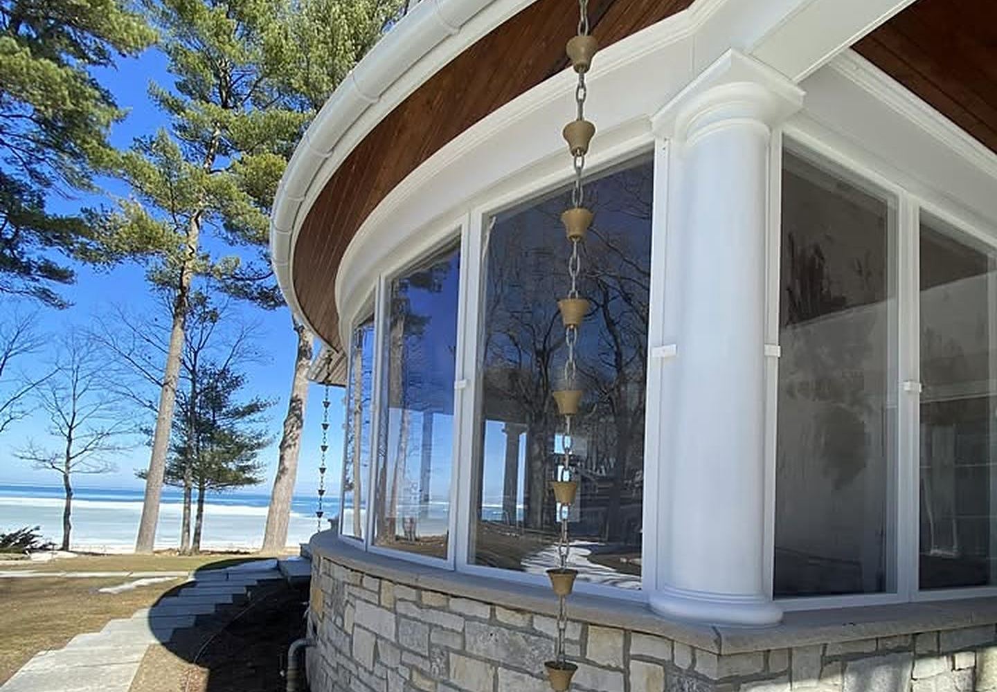 A copper rain chain hanging from a white half-round gutter on a stone and glass lakefront home
